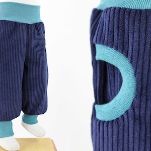 Pode incluir: Calças de veludo cotelê azul com detalhes turquesa. As calças têm um design único com um círculo turquesa recortado na lateral. As calças têm uma cintura e punhos turquesa.