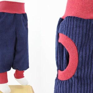 Pode incluir: Calça de veludo cotelê azul com detalhes em vermelho e um design único com um círculo vermelho recortado na lateral. A calça tem uma cintura vermelha e punhos vermelhos.