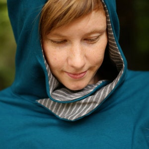 Op de afbeelding: Een vrouw draagt een teal blauwe hoodie met een grijze en witte gestreepte rand rond de capuchon.