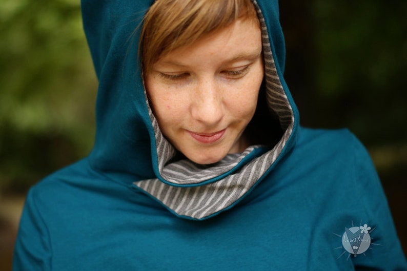 Op de afbeelding: Een vrouw draagt een teal blauwe hoodie met een grijze en witte gestreepte rand rond de capuchon.