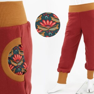 Op de afbeelding: Roestkleurige joggingbroek met een mosterdgele tailleband, manchetten en zakafwerking. De zakken hebben een bloemenpatroon in teal, rood en goud. De broek heeft een relaxte pasvorm en wordt op een mannequin getoond.