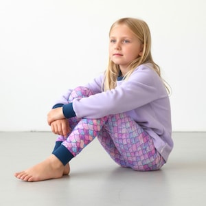 Könnte beinhalten: Ein Kind trägt einen lavendelfarbenen Fleece-Sweatshirt mit marineblauen Bündchen und eine Leggings mit Meerjungfrauenschuppen. Die Leggings haben ein farbenfrohes Muster aus rosa, lila und blauen Schuppen. Das Kind sitzt auf dem Boden.