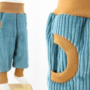 Pode incluir: Calças de veludo cotelê azul com detalhes em veludo cotelê castanho. As calças têm um bolso único em forma de lua crescente na frente.