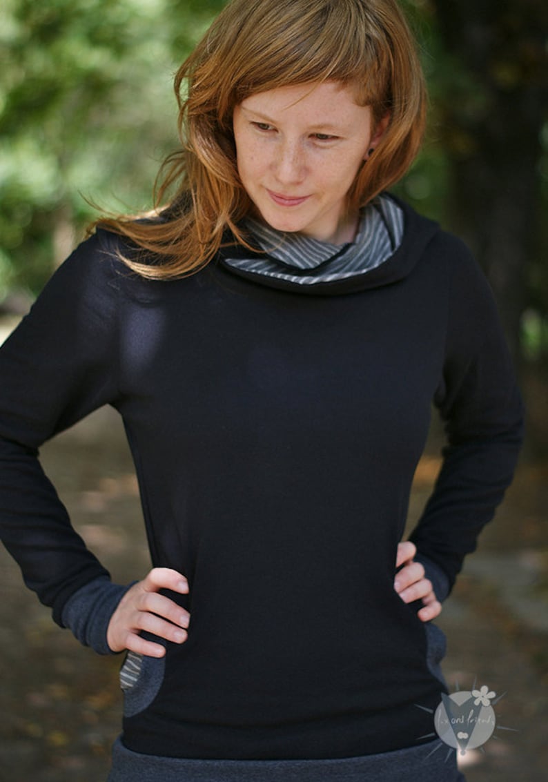May include: A woman wearing a black hooded sweatshirt with a striped cowl neck. The sweatshirt has long sleeves and a grey waistband.