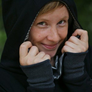May include: A person wearing a black hooded sweatshirt with a striped lining. The hood is pulled up and the person is smiling.