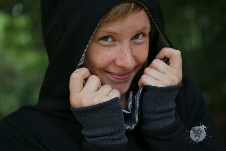 May include: A person wearing a black hooded sweatshirt with a striped lining. The hood is pulled up and the person is smiling.
