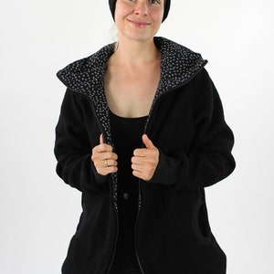 May include: A black fleece jacket with a zipper closure and a hood. The jacket has a white and black patterned lining. The model is wearing black jeans and a black headband.
