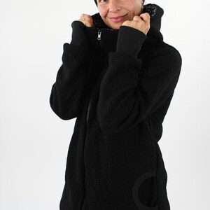May include: A black fleece jacket with a zipper closure and a hood. The jacket has a large, round pocket on the left side. The model is wearing black pants and a black headband.