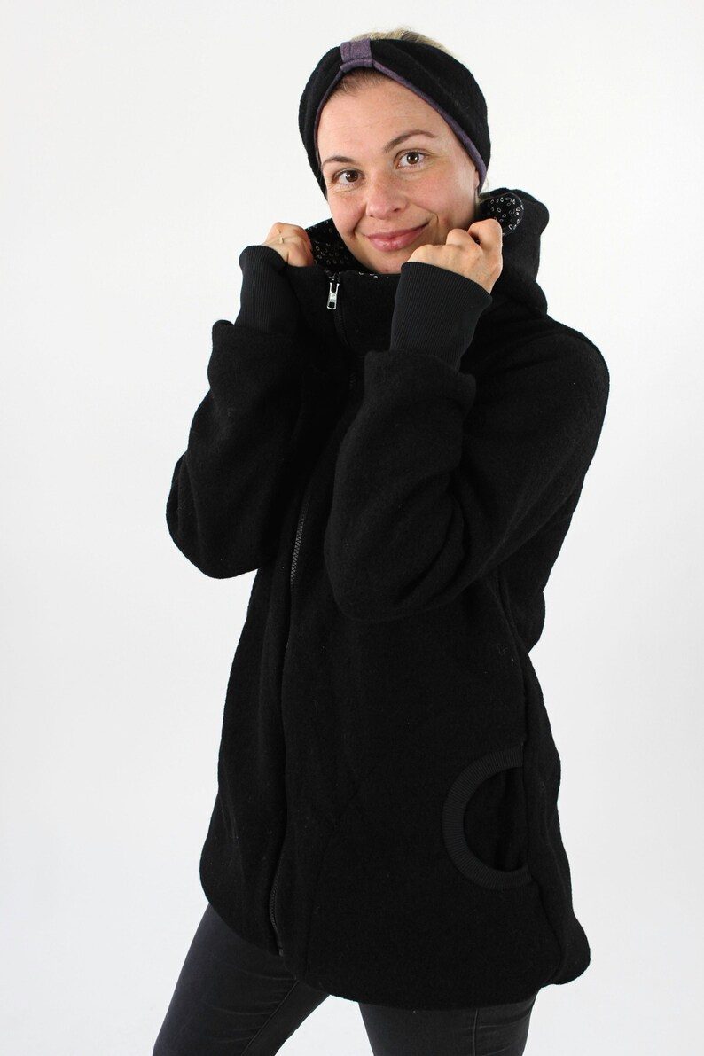 May include: A black fleece jacket with a zipper closure and a hood. The jacket has a large, round pocket on the left side. The model is wearing black pants and a black headband.