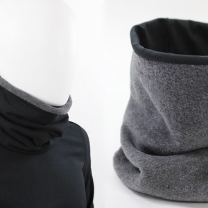 Könnte beinhalten: Ein grauer Fleece-Halswärmer mit schwarzem Fleece-Futter. Der Halswärmer ist gefaltet und auf einer weißen Oberfläche abgelegt.