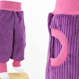 Pode incluir: Calça de veludo cotelê roxo com cintura rosa e acabamento rosa nos bolsos. A calça tem um design único com um grande recorte arredondado na lateral de cada bolso.