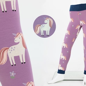 Könnte beinhalten: Lavendelfarbene Leggings mit einem sich wiederholenden Muster aus weißen Einhörnern mit rosa Mähnen und Schweifen sowie kleinen Sternakzenten. Die Leggings haben einen dunkelblauen Bund und Bündchen. Eine kreisförmige Grafik zeigt ein Einhorn.