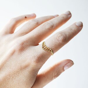 A gold toned textured chevron shaped ring with open ovals within the ring. The largest oval at the center and they get smaller as they get towards the sides of the ring. Shown on the pointer finger of a hand.