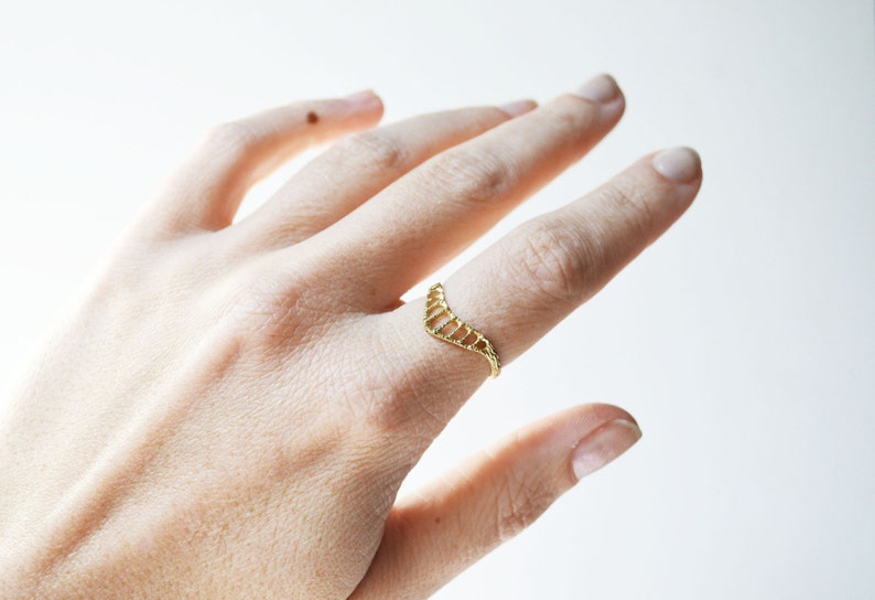 A gold toned textured chevron shaped ring with open ovals within the ring. The largest oval at the center and they get smaller as they get towards the sides of the ring. Shown on the pointer finger of a hand.