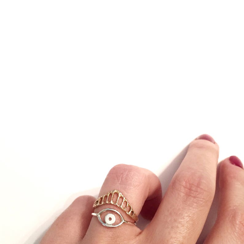 A silver textured chevron shaped ring with open ovals within the ring. The largest oval at the center and they get smaller as they get towards the sides of the ring. Shown stacked above another eye shaped ring.