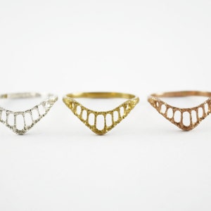 Three chevron shaped rings with open ovals within the ring. The largest oval at the center and they get smaller as they get towards the sides of the ring. Three shown in a line from left, silver, yellow brass and 14k rose gold.