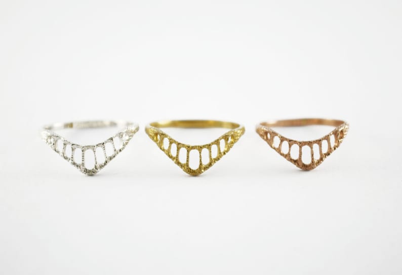 Three chevron shaped rings with open ovals within the ring. The largest oval at the center and they get smaller as they get towards the sides of the ring. Three shown in a line from left, silver, yellow brass and 14k rose gold.