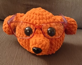 May include: A crocheted orange bear wearing pink glasses. The bear has black eyes and a black nose.