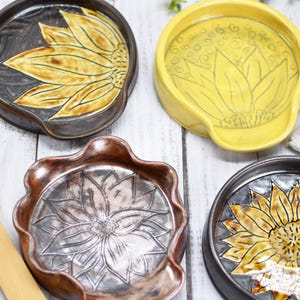 May include: Four ceramic spoon rests with sunflower and floral motifs in yellow, brown, and metallic copper glazes. Two bamboo kitchen utensils, a silver spoon, a white coffee pod, and a green tea bag are arranged alongside the ceramic dishes.