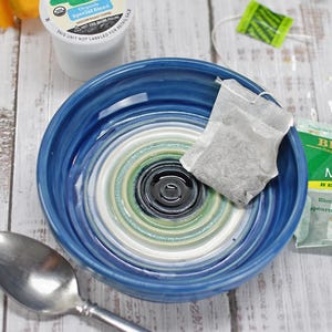 May include: A blue ceramic bowl with concentric rings in white, green, and black at the center. The bowl contains a tea bag. Nearby are a silver spoon, a coffee pod labeled Coffee, and a green Bigelow Mint Medley herbal tea bag packet.