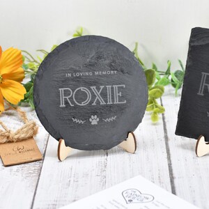 May include: Two slate coasters, one round and one square, each engraved with "IN LOVING MEMORY" and the name "ROXIE", along with a paw print and floral accents. They are displayed on wooden stands, with flowers in the background.