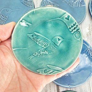 May include: A round, teal ceramic coaster featuring an embossed hummingbird design with intricate patterns on its wings and a small heart shape on its head. The surface has a glossy finish, and the edges are smooth and slightly raised.