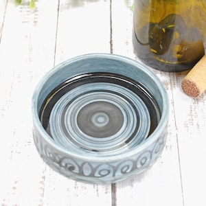 May include: A round, blue ceramic coaster with a concentric circle design in shades of gray and black. The coaster has a decorative pattern around the edge. A wine bottle and cork are also visible.