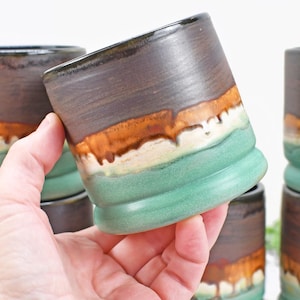May include: Handmade ceramic tumblers with a unique design. The cups feature a dark brown top, transitioning to a band of orange and white, and a turquoise base. The tumblers are stacked, showcasing their layered color scheme and textured finish.