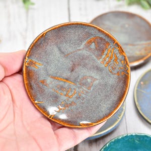 May include: Handmade ceramic coasters in various colors, including gray, blue, and teal. Each coaster features a unique design in shades of brown and orange. The coasters are round and appear to be approximately 4 inches in diameter.