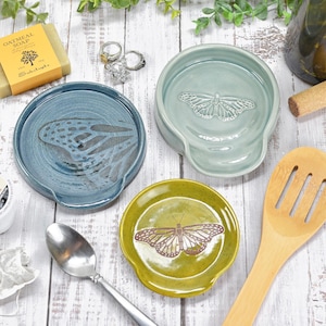 May include: A collection of ceramic spoon rests in blue, green, and yellow, each with a butterfly design. Also includes a wooden spoon, soap, coffee pod, tea bag, rings, and a wine bottle with a cork.
