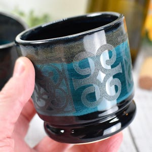 May include: A pair of ceramic shot glasses with a black base, teal and gray midsection, and a dark blue rim. Each glass features a silver, stylized design. One glass is held in a hand, with a second glass visible in the background.
