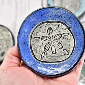 May include: A set of four blue and white ceramic coasters with a sand dollar design. The coasters are hand-painted and have a speckled glaze.