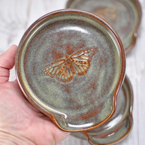 May include: A round ceramic spoon rest featuring a mottled sage green and rustic brown glaze with a raised, detailed butterfly motif centered in the basin. The edges are smooth and curved, with a distinct indentation for resting a kitchen utensil.