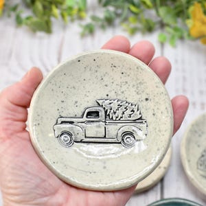 May include: A small, speckled, off-white ceramic dish featuring a raised relief of a vintage pickup truck carrying a Christmas tree. The truck is detailed in silver, and the dish has a slightly rustic, handmade appearance. The dish is round and has a shallow bowl shape.