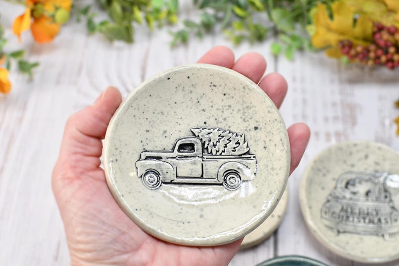 May include: A small, speckled, off-white ceramic dish featuring a raised relief of a vintage pickup truck carrying a Christmas tree. The truck is detailed in silver, and the dish has a slightly rustic, handmade appearance. The dish is round and has a shallow bowl shape.