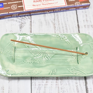May include: A rectangular light green ceramic incense holder featuring embossed hummingbird and floral motifs. The tray has a glossy finish with a small hole at one end for holding a single brown incense stick, which is positioned horizontally.