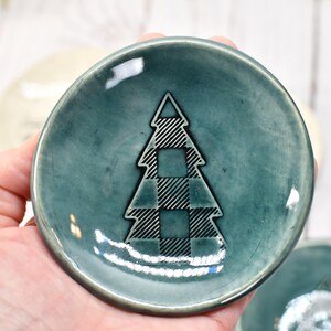 May include: A small, teal-colored ceramic dish with a plaid Christmas tree design in the center. The dish is round and has a slightly raised edge. Other similar dishes are visible in the background.