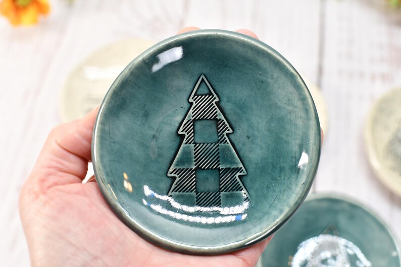 May include: A small, teal-colored ceramic dish with a plaid Christmas tree design in the center. The dish is round and has a slightly raised edge. Other similar dishes are visible in the background.