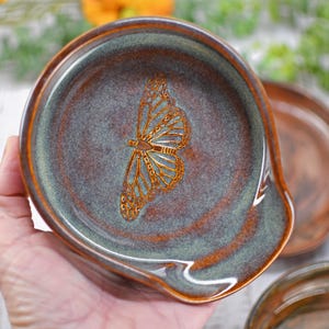 May include: A round ceramic spoon rest featuring a glossy glaze in shades of deep brown and muted green. The center of the spoon rest displays a detailed, etched golden-brown butterfly wing pattern. The edges are smooth with a curved handle rest.