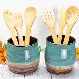 May include: Two teal ceramic utensil holders with brown and white accents. Each holder contains a variety of wooden kitchen utensils, including spoons, spatulas, and a slotted spoon.