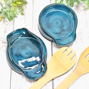 May include: Two teal ceramic spoon rests with a glossy finish, alongside two bamboo cooking utensils. The spoon rests have a unique shape and are placed on a white wooden surface, with greenery and a flower in the background.