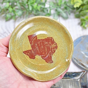 May include: A round, ceramic spoon rest with a speckled, olive-green glaze. The center features a distressed, reddish-brown outline of the state of Texas. The spoon rest is held in a hand, with other ceramic pieces in the background.