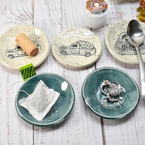 May include: A collection of small, ceramic dishes and trays. Some are off-white with black truck illustrations, a cork, and the text "Merry Christmas". Others are teal-colored, holding a tea bag and rings. A silver spoon rests on one dish.