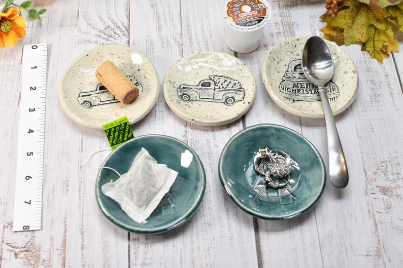 May include: A collection of small, ceramic dishes and trays. Some are off-white with black truck illustrations, a cork, and the text "Merry Christmas". Others are teal-colored, holding a tea bag and rings. A silver spoon rests on one dish.