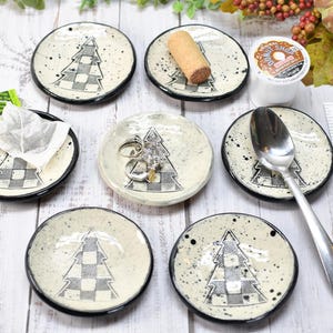 May include: A collection of small, round ceramic dishes with a black rim and a speckled, off-white surface. Each dish features a black and white checkered Christmas tree design. Some dishes hold items like a tea bag, rings, and a spoon.