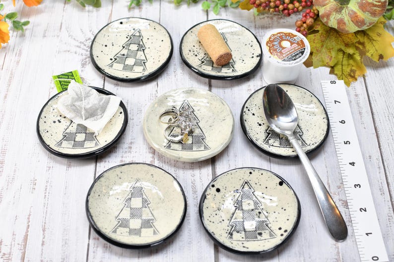 May include: A collection of small, round ceramic dishes with a black rim and a speckled, off-white surface. Each dish features a black and white checkered Christmas tree design. Some dishes hold items like a tea bag, rings, and a spoon.