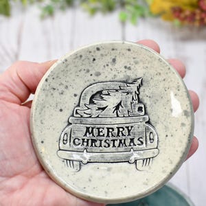 May include: A round, speckled ceramic coaster with a raised design of a car carrying a Christmas tree. The words "MERRY CHRISTMAS" are displayed on the car. The coaster is light gray with dark gray speckles.