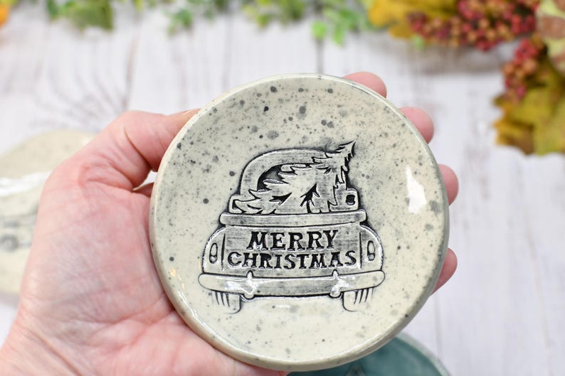 May include: A round, speckled ceramic coaster with a raised design of a car carrying a Christmas tree. The words "MERRY CHRISTMAS" are displayed on the car. The coaster is light gray with dark gray speckles.