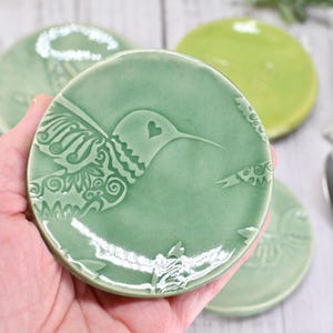 May include: A set of four ceramic coasters in shades of green. Each coaster features a detailed hummingbird design with a heart accent. The coasters are round and appear to be handmade, adding a touch of nature-inspired decor.