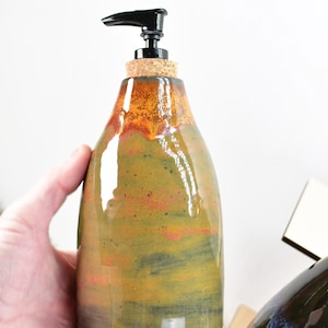 May include: A ceramic soap dispenser with a black pump and a cork top. The dispenser is hand-painted with a green, orange, and brown glaze.